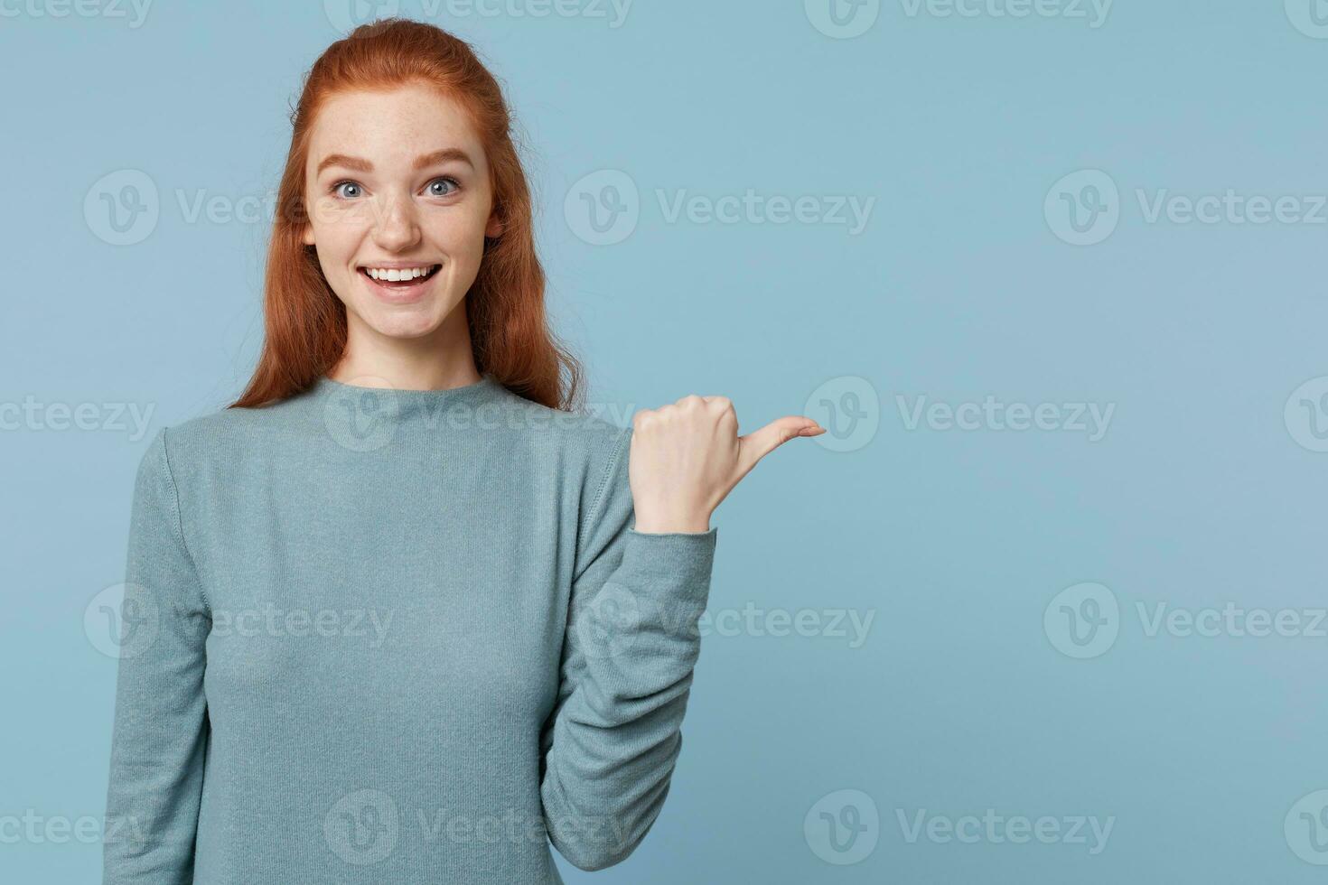 A snapshot of a young red-haired girl who smiles and happily shows with her thumb in the direction of the empty copy space, isolated on a blue background. photo