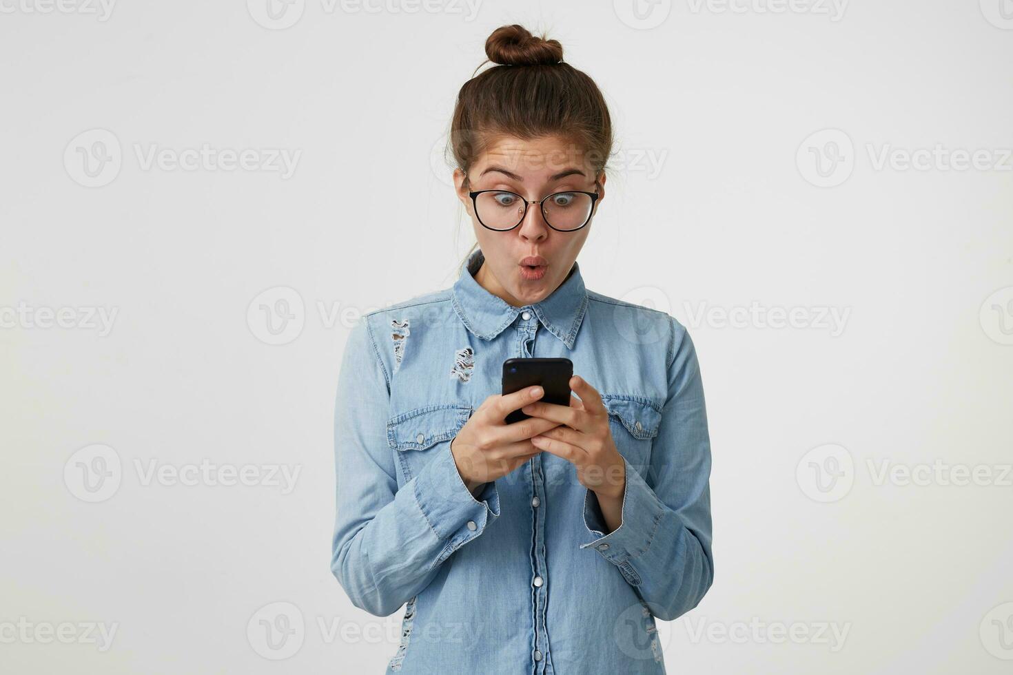 Young woman in glasses is looking at, holding in hands a smartphone, her eyes wide open and rounded mouth in surprisement, amazement. Shocked, read the sensation, staggering news. photo