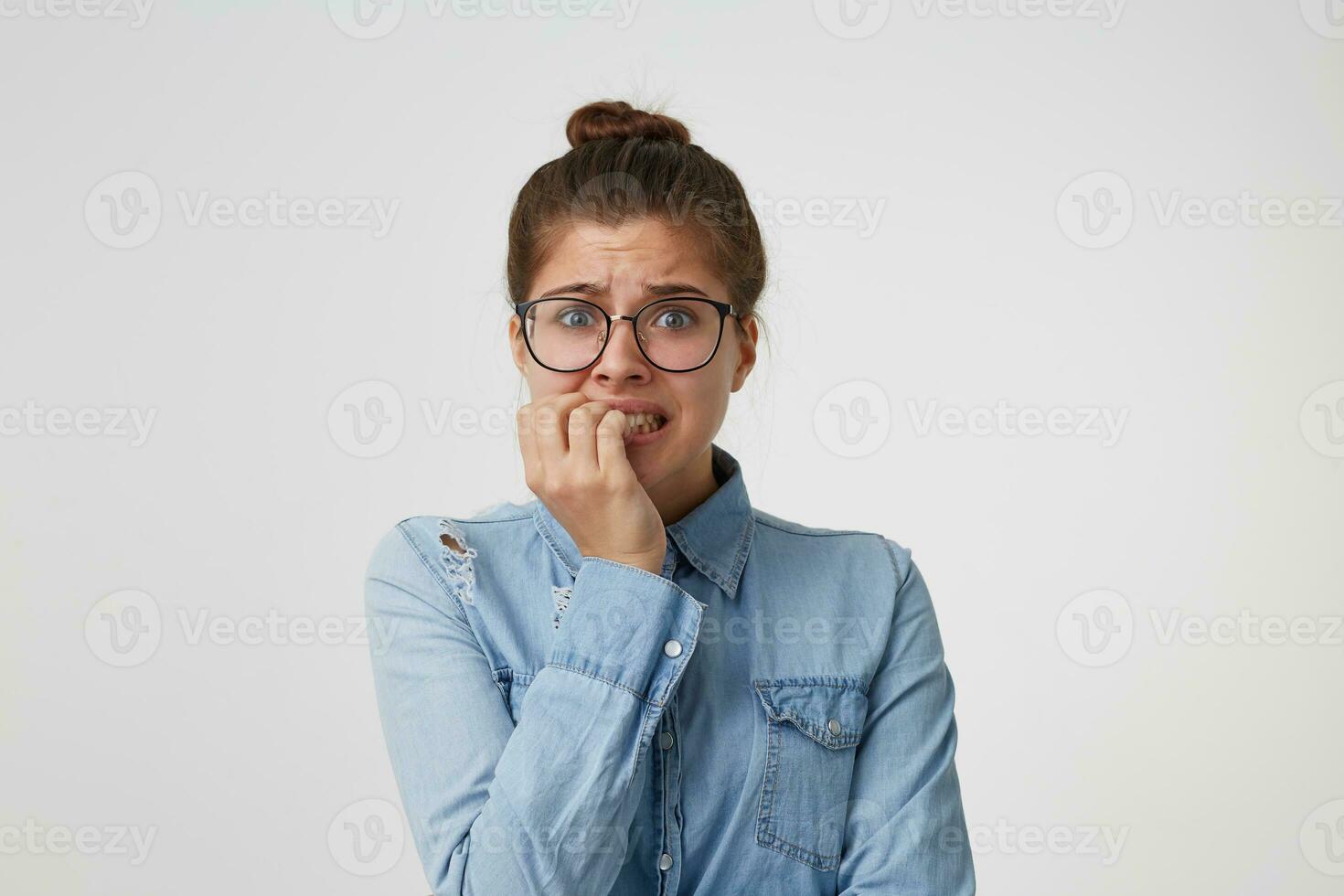 Portrait of a girl student in glasses, dressed in a fashionable denim shirt, worries about something, nervous,bites her nails, waiting for the exam results isolated on white background photo