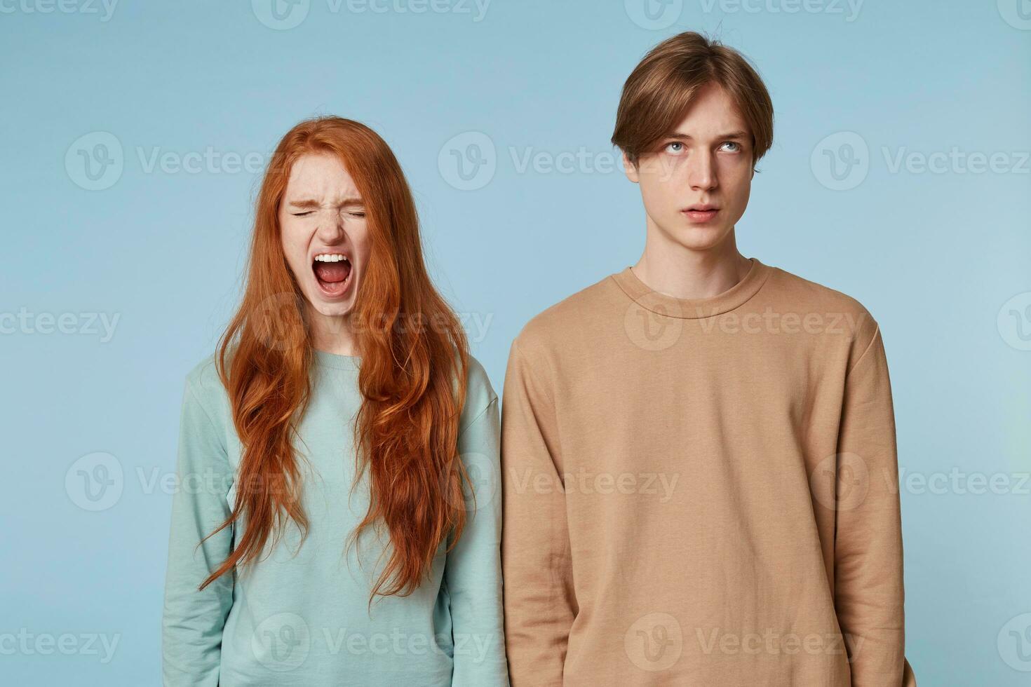 Couple relationship. A girl with red long hair stands with eyes closed opening her mouth wide as if screaming, the guy next to her is rolling his eyes up tired of listening to the cries, of quarreling photo