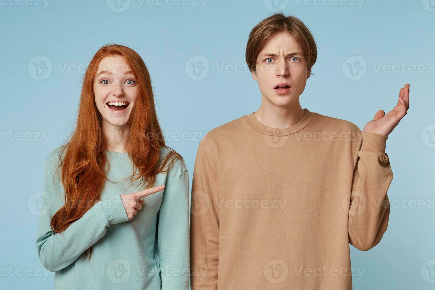 Couple,girl with long red hair laughs shows with her index finger at the guy who stands with a bewildered, confused, puzzled expression,hand raised up,isolated on blue background photo