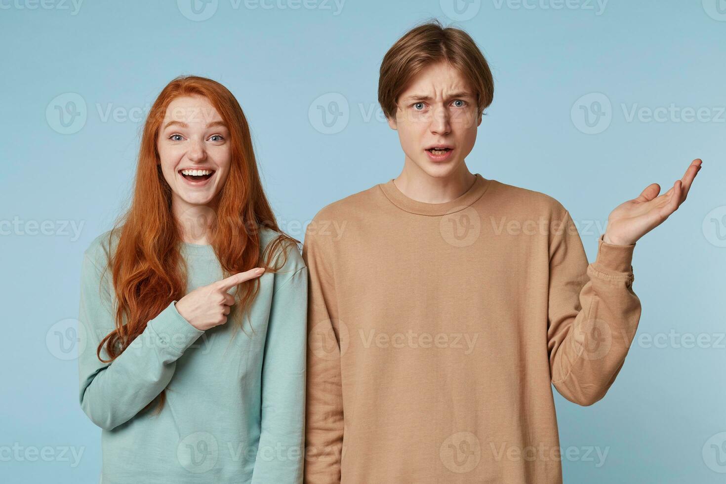 Couple,girl with long red hair laughs shows with her index finger at the guy who is standing with an uncomprehending frown expression outraged,something says,hand raised up,isolated on blue background photo