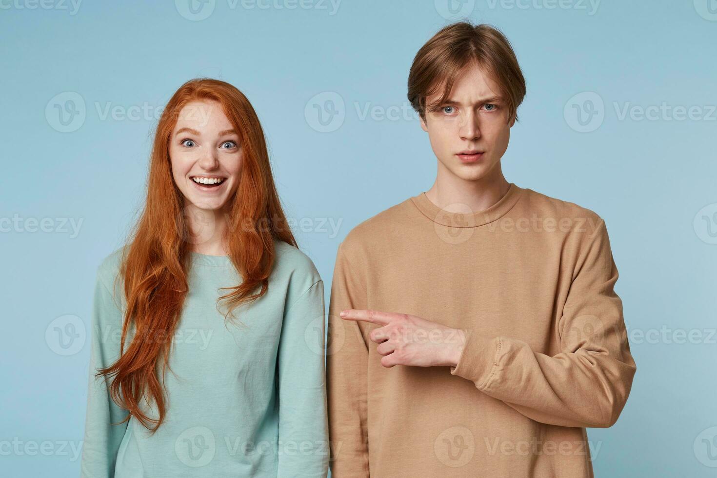 Close up couple, girl smiles joyful happy with wide open eyes and a smile,long red hair, the guy is not in the spirit displeased points his index finger to his girlfriend,isolated on a blue background photo