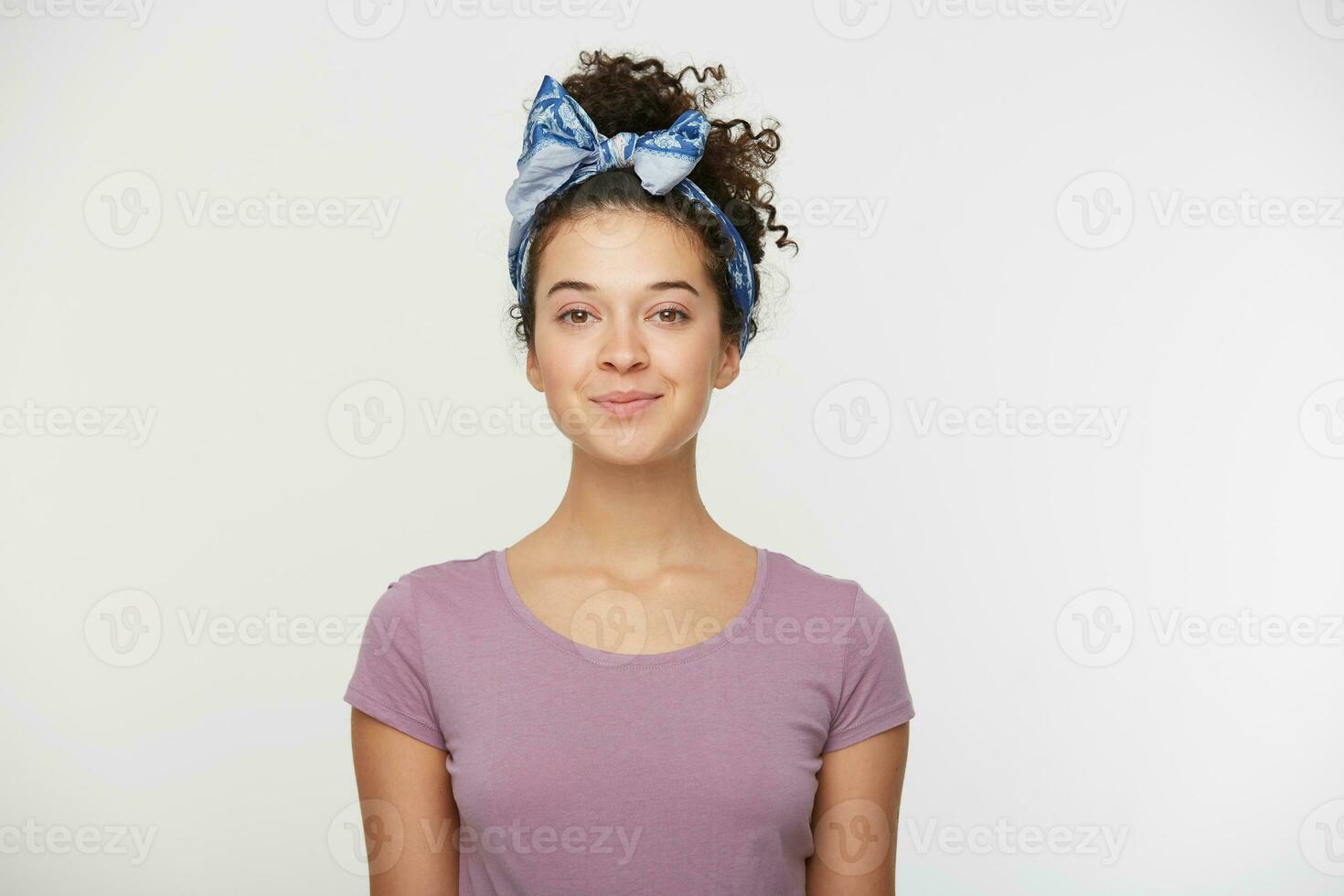 Happy female with positive expression, dressed in casual t shirt and stylish headband, poses against white background, looking at the camera with a playful smile. People and emotions photo