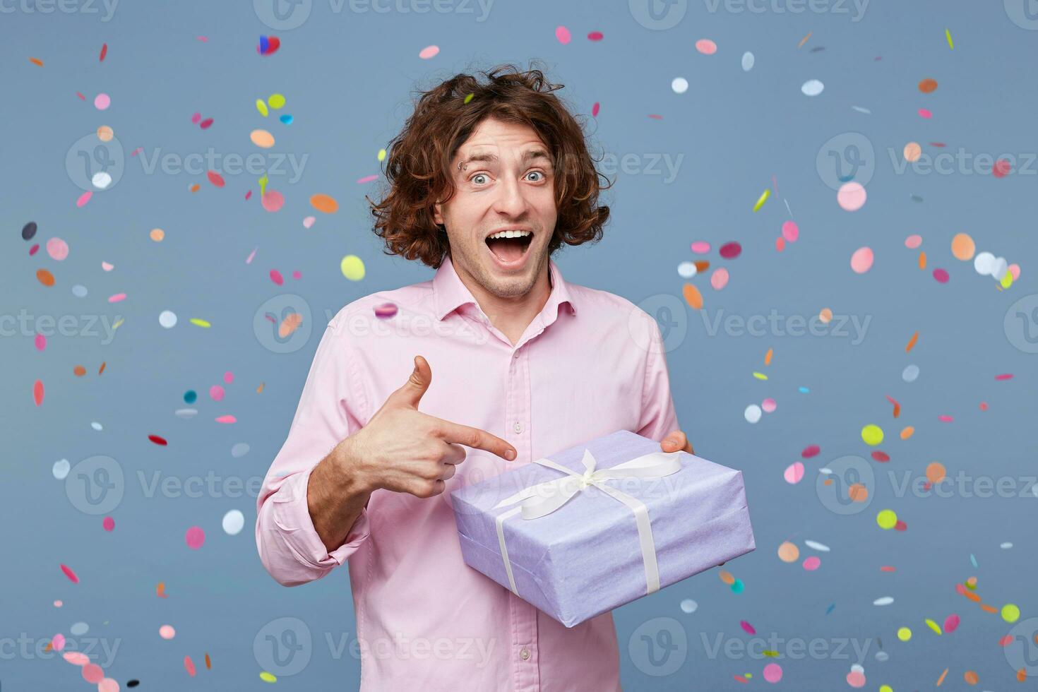 Exited happy birthday guy got a surprise party, posing for picture, holding box with present and pointing on it with index finger, confetti falling down. People, joy, fun and happiness concept. photo
