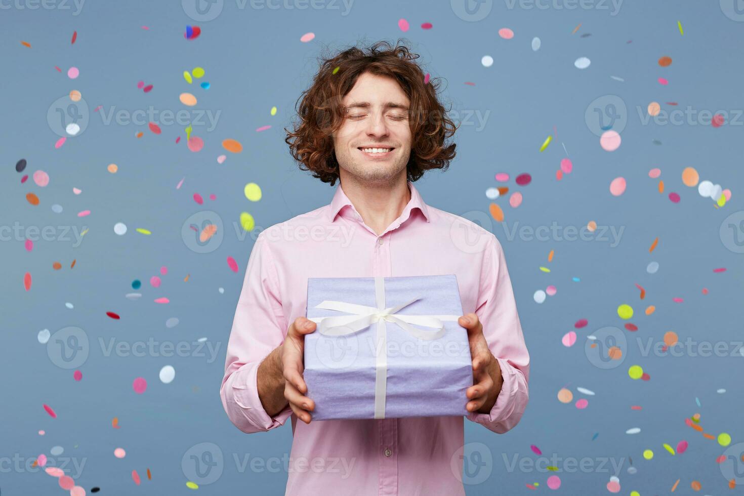 People, joy, fun and happiness concept. Relaxed happy birthday guy with eyes closed waiting for a surprise, posing for picture, holding box with present and with confetti falling down photo