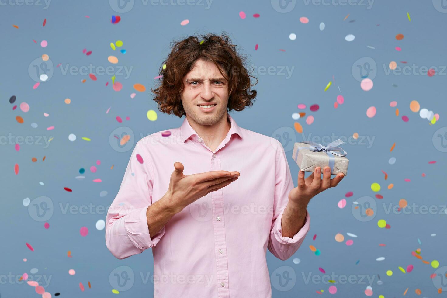 The birthday man was congratulated, he was given a gift, he stands displeased with a puzzled expression on his face and is pointing on a small box with a present in his hand. photo
