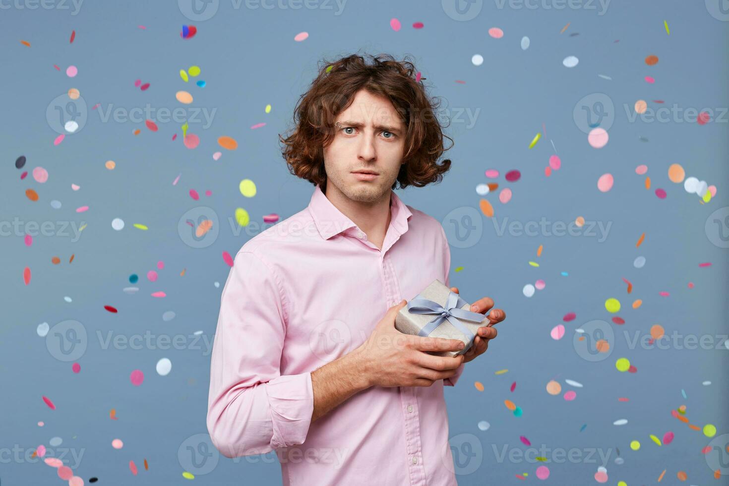 Guy posing with a gift box looks as if he is afraid that someone will take it away, does not want to part with it, holds present tightly in his hands, against the background of falling confetti. photo