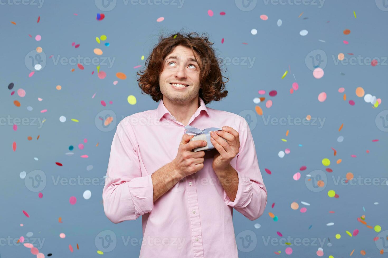 The guy holds a gift that he was handed, looks away imagining what lies in the box, dreams of something, confetti fall down isolated on a blue background photo
