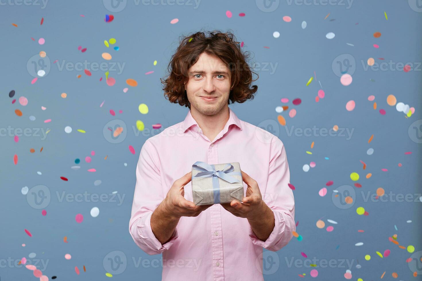 Portrait of a guy who gives a small gift, tied with a blue ribbon, is going to give a modest gift from the heart, looking at the camera, confetti falling down. People, joy, fun and happiness concept. photo