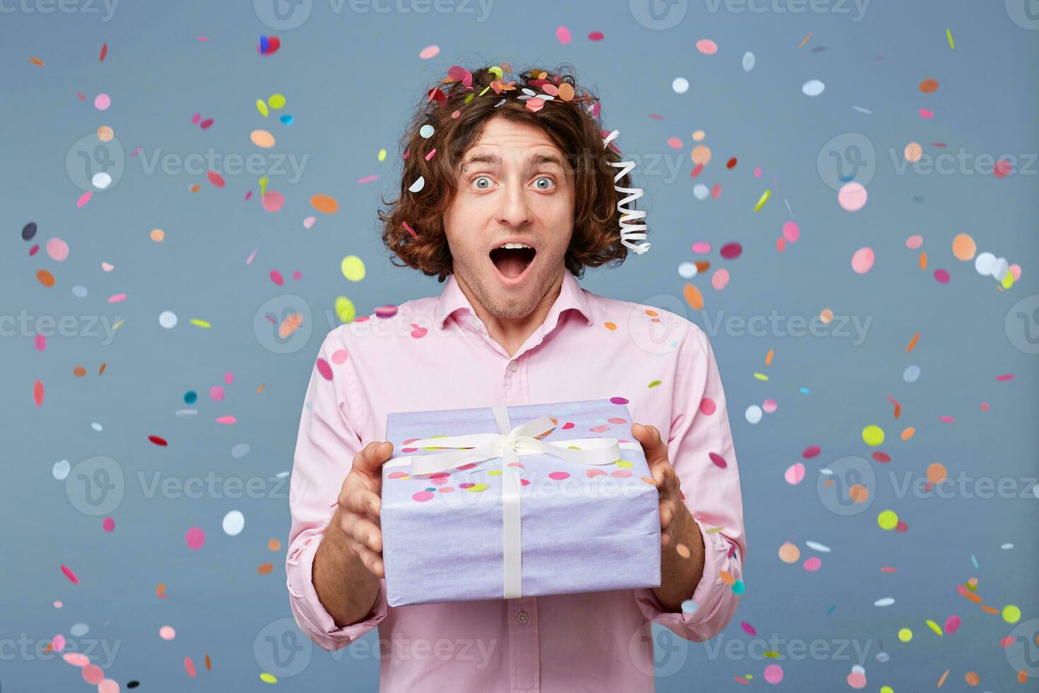 Cheerful happy young male man with a decorative gift isolated on blue background. Man is receiving gift box. He is standing and looking at camera with surprise. photo