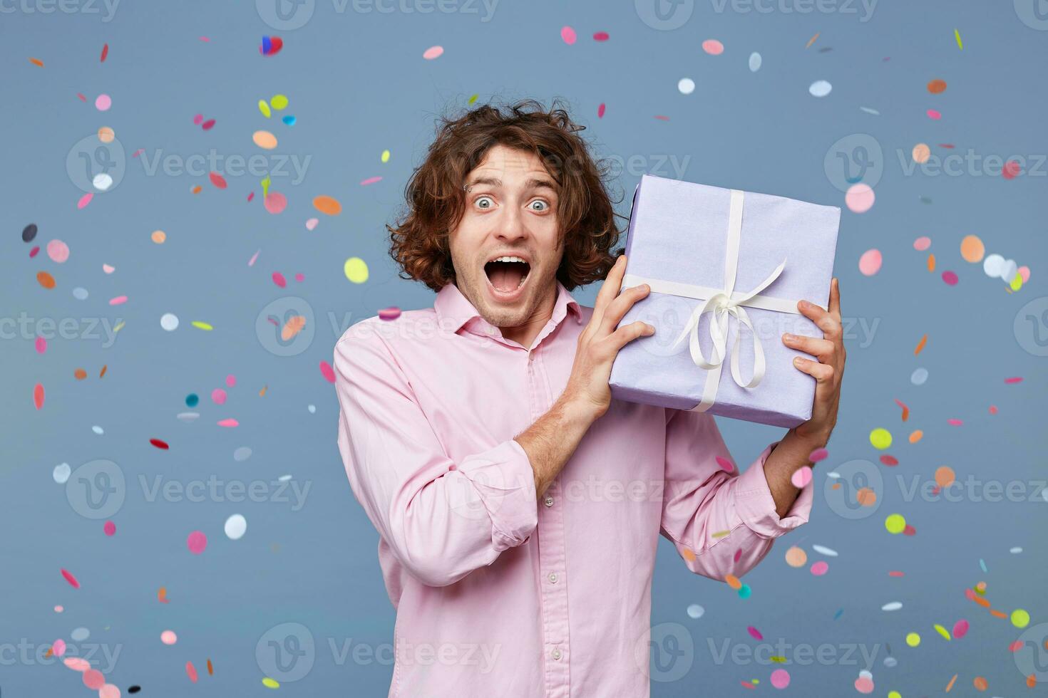 Joy, great fun, happiness, portrait of a birthday man received a gift box, happy photographed around falling confetti on a blue background. The man was congratulated handed a gift, he delighted. photo