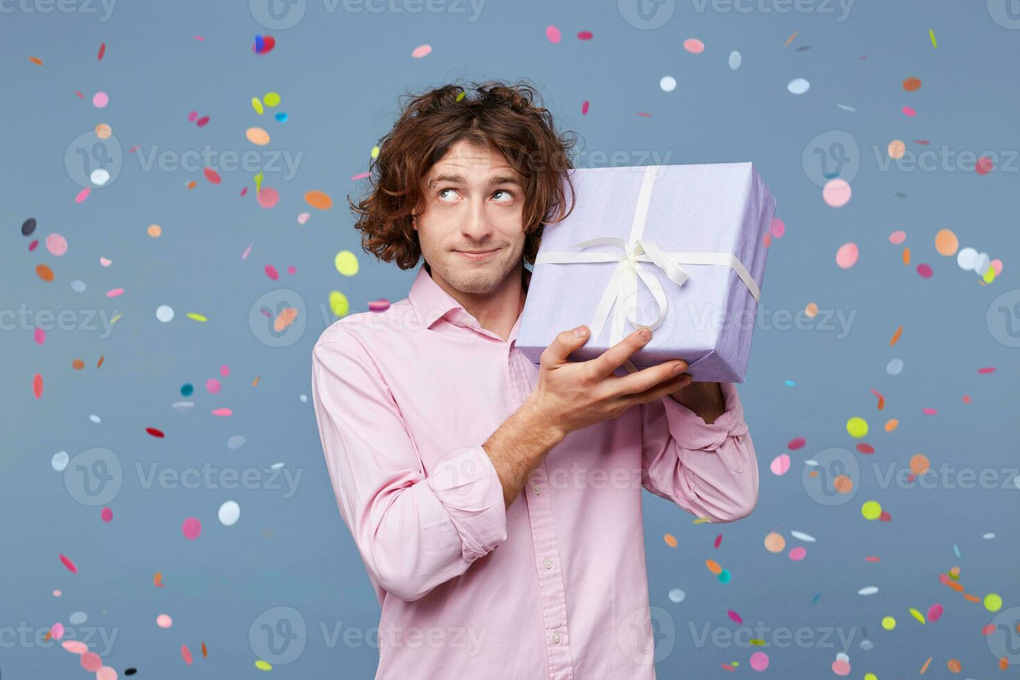 The birthday man received a box as a gift, holds it in his hands near his head, looks away wondering guessing about what's inside, confetti falling down. People, joy, fun and happiness concept. photo