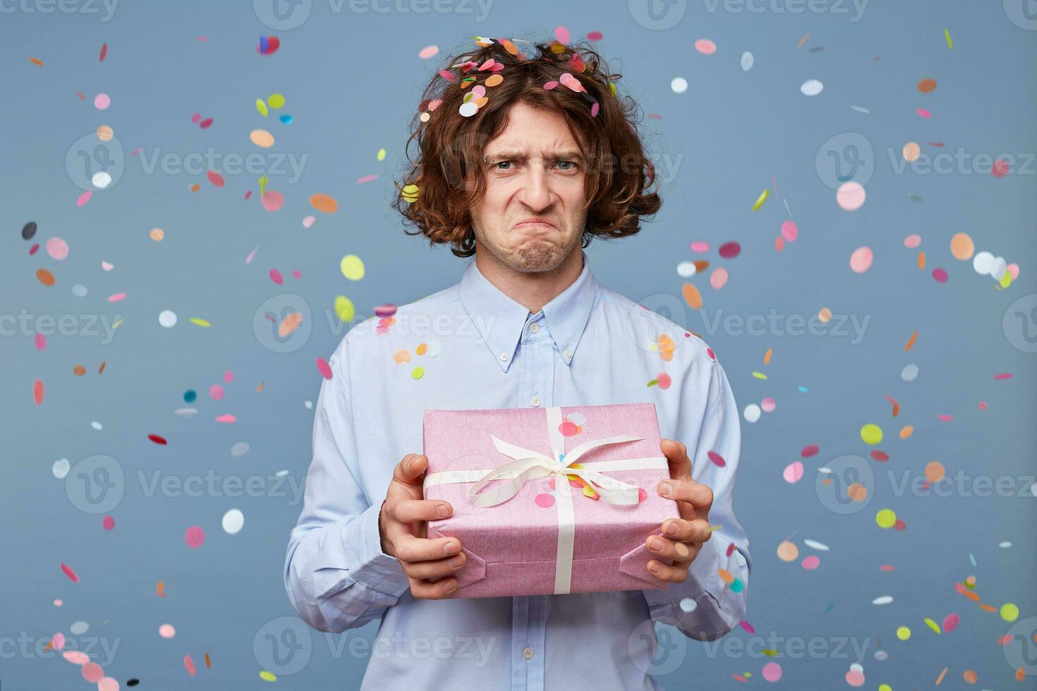 Portrait of a guy holding a present box with a frustrated expression, pursed his lips, dissatisfied, upset, offended, looking into the camera standing against the blue background of falling confetti. photo