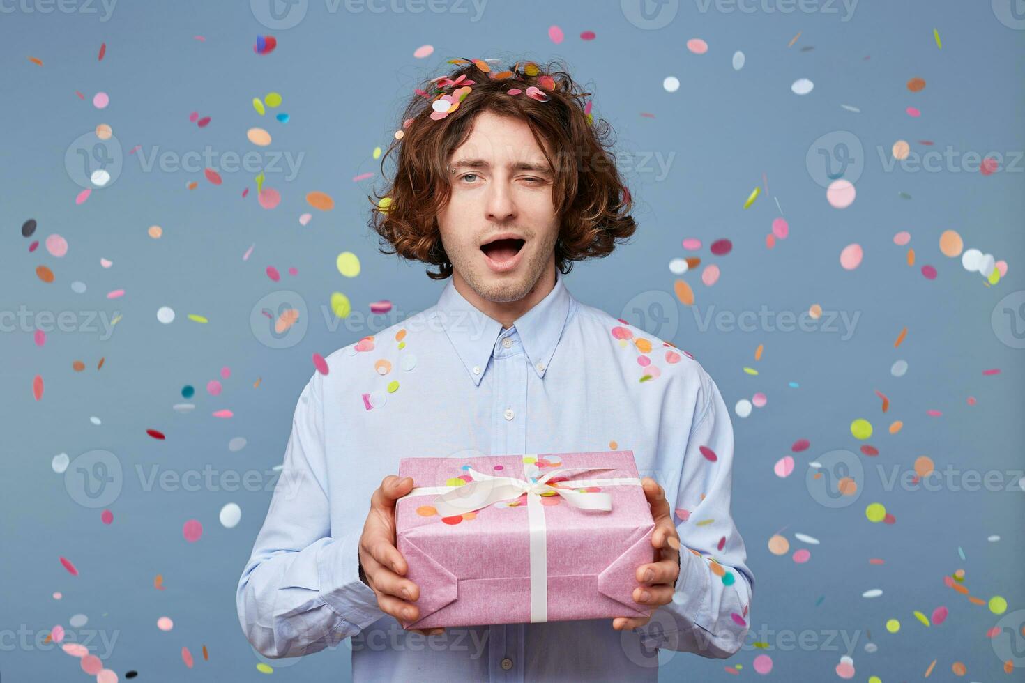 Man holding a decorated gift in a big box, presents it, winkles, in a playful mood, confetti falling down on a blue background. photo