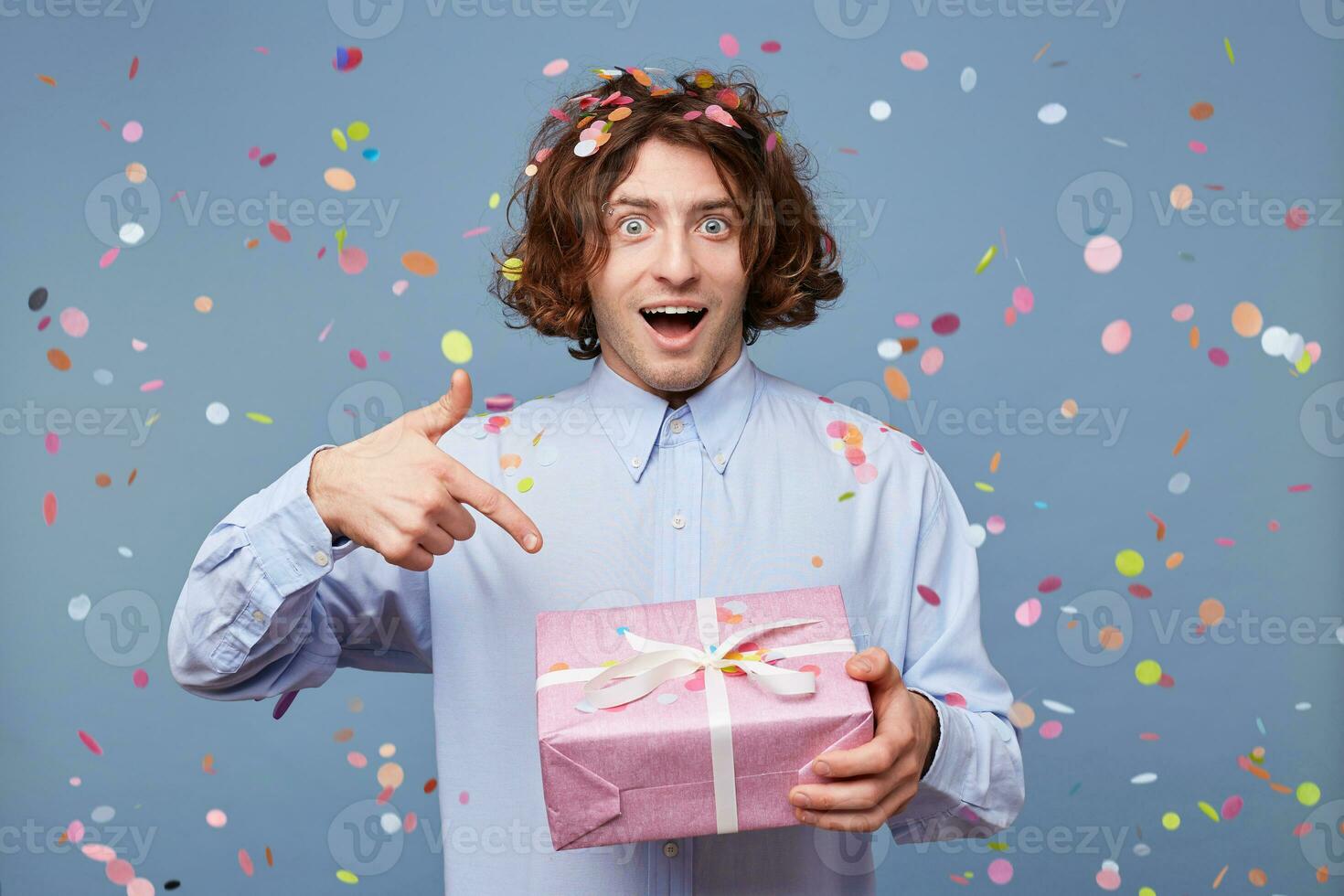 Festive, lively birthday guy got posing for picture, holding box with present and pointing on it with index finger, confetti falling down. People, joy, fun and happiness concept. photo