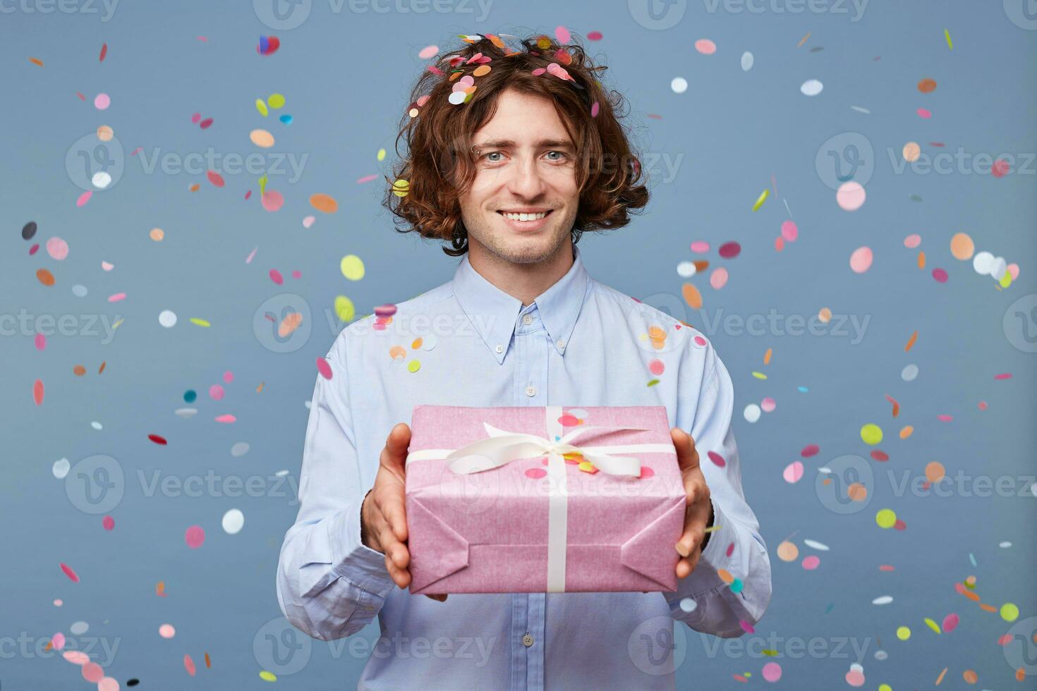 Close up of a man who gives a gift in a pink box with white ribbon, is going to give a modest gift from the heart, looking at the camera, confetti falling down. People, joy, fun and happiness concept. photo