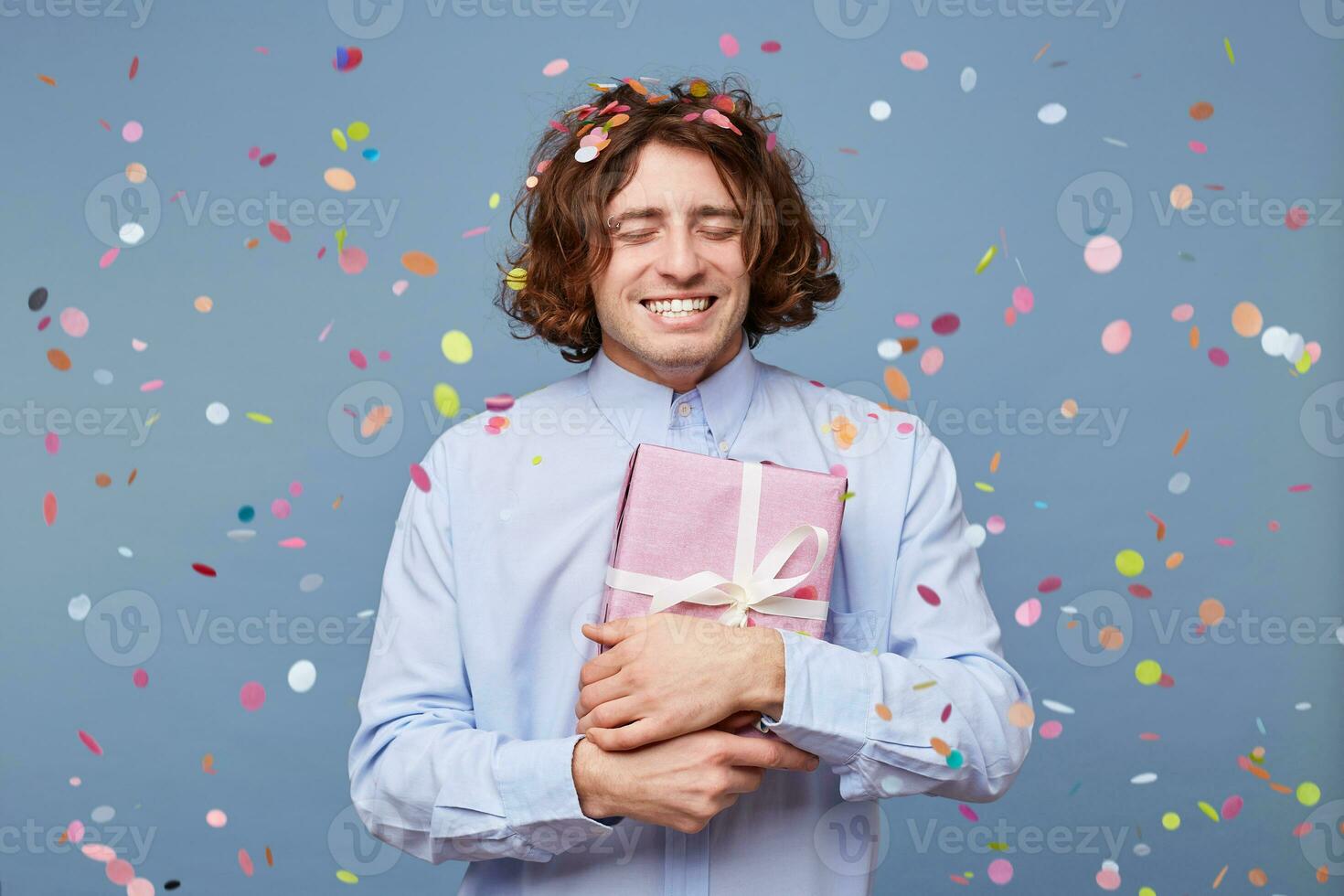 cumpleaños chico con ojos cerrado, prensas el regalo caja atado con un blanco cinta a él mismo, es agradecido feliz, disfruta momento, papel picado caídas en él, se encuentra aislado en un azul fondo, siente felicidad foto