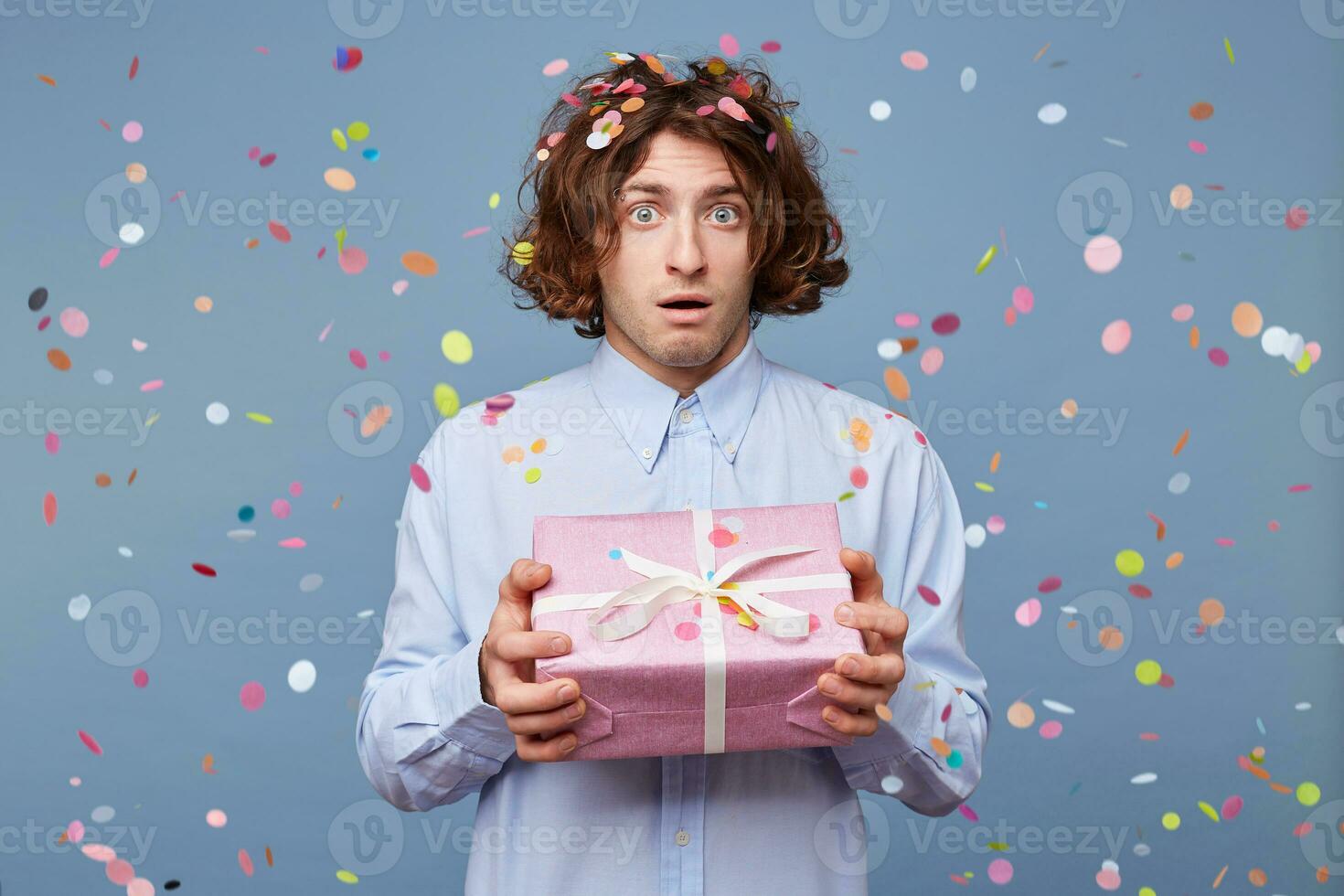 Indoor shot of amazed man who received a new year gift, keeps mouth wide opened, stares at camera, says omg or wow. Pleasant surprised male, standing against the blue background of falling confetti. photo