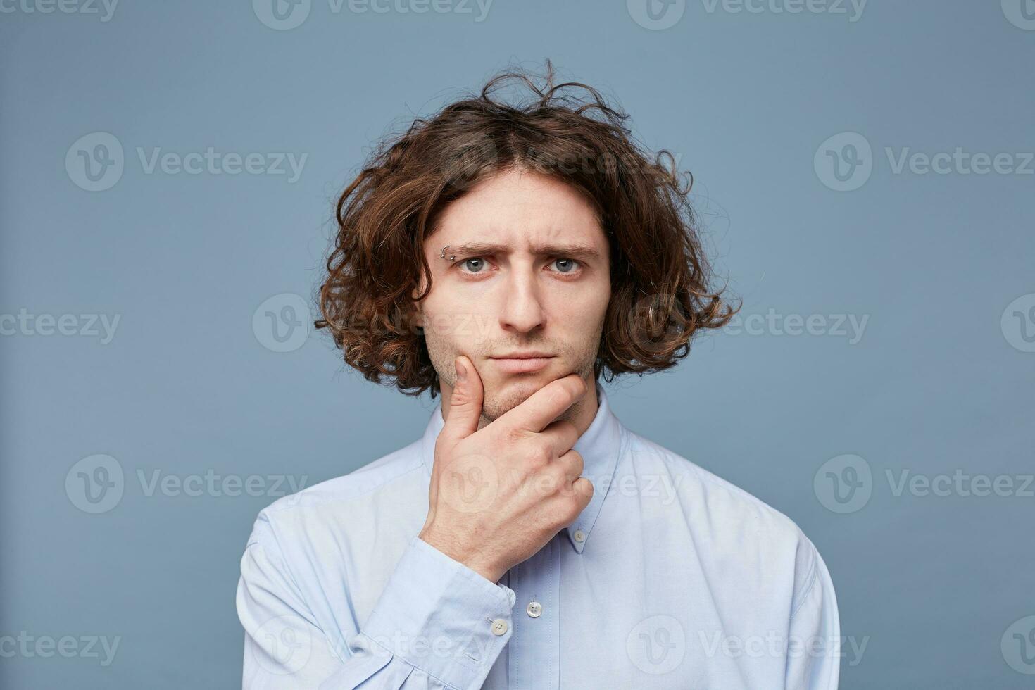 Portrait of a pensive young man dressed in shirt holding hand on his chin isolated over blue background. Creative and artistic male gazing thoughtfully at camera, thinking or making up ideas photo