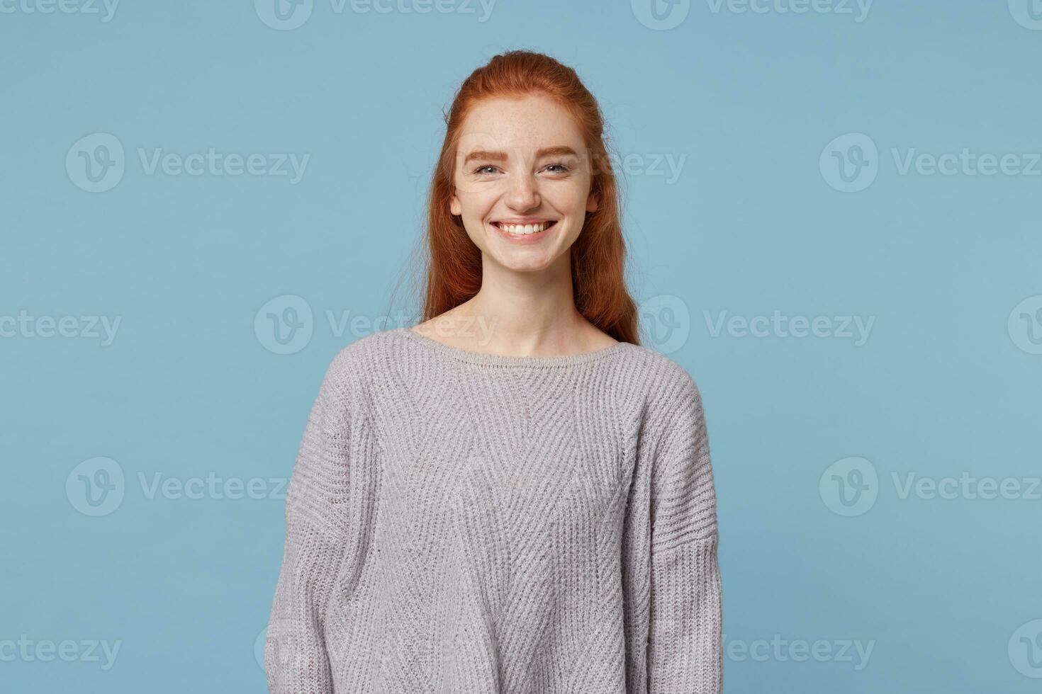 Beautiful attractive young red-haired girl cute openly cheerfully smiling, showing her teeth, wearing an oversized warm sweater, isolated on a blue background photo