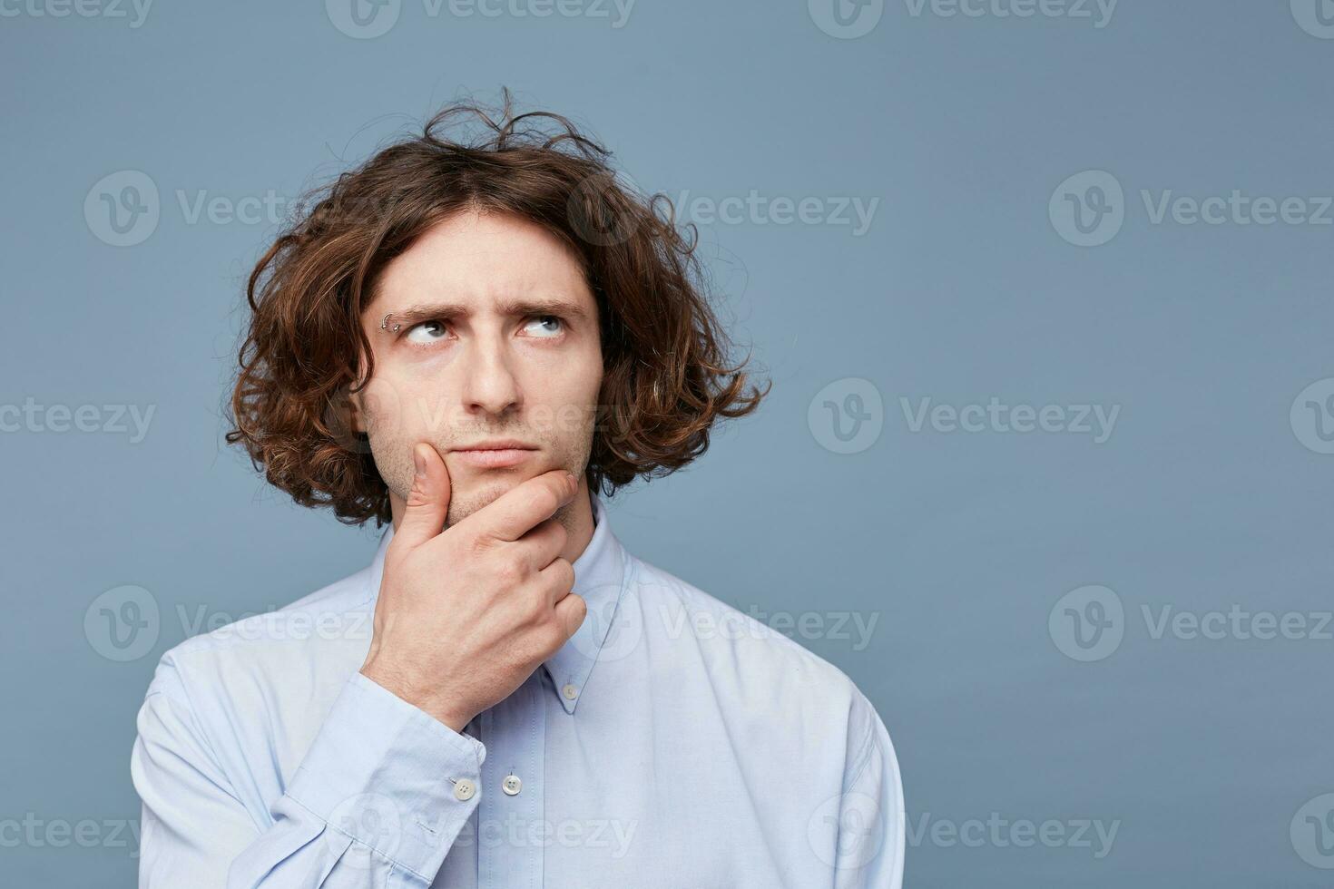 Portrait of suspicious pensive young male in casual shirt touching face while thinking over something, trying to come up with solution, having perplexed and puzzled expression over blue background photo