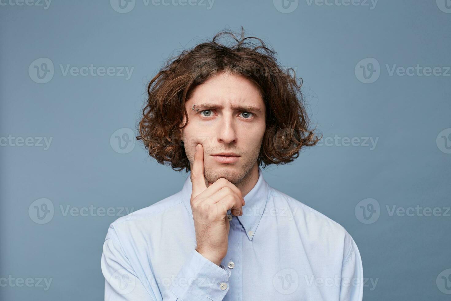 Thoughts and doubts concept. Man with long messy hair thoughtful face, holding fingers on cheek and chin, gazing camera. Guy with untidy hairstyle has doubts, copy space, blue background. photo