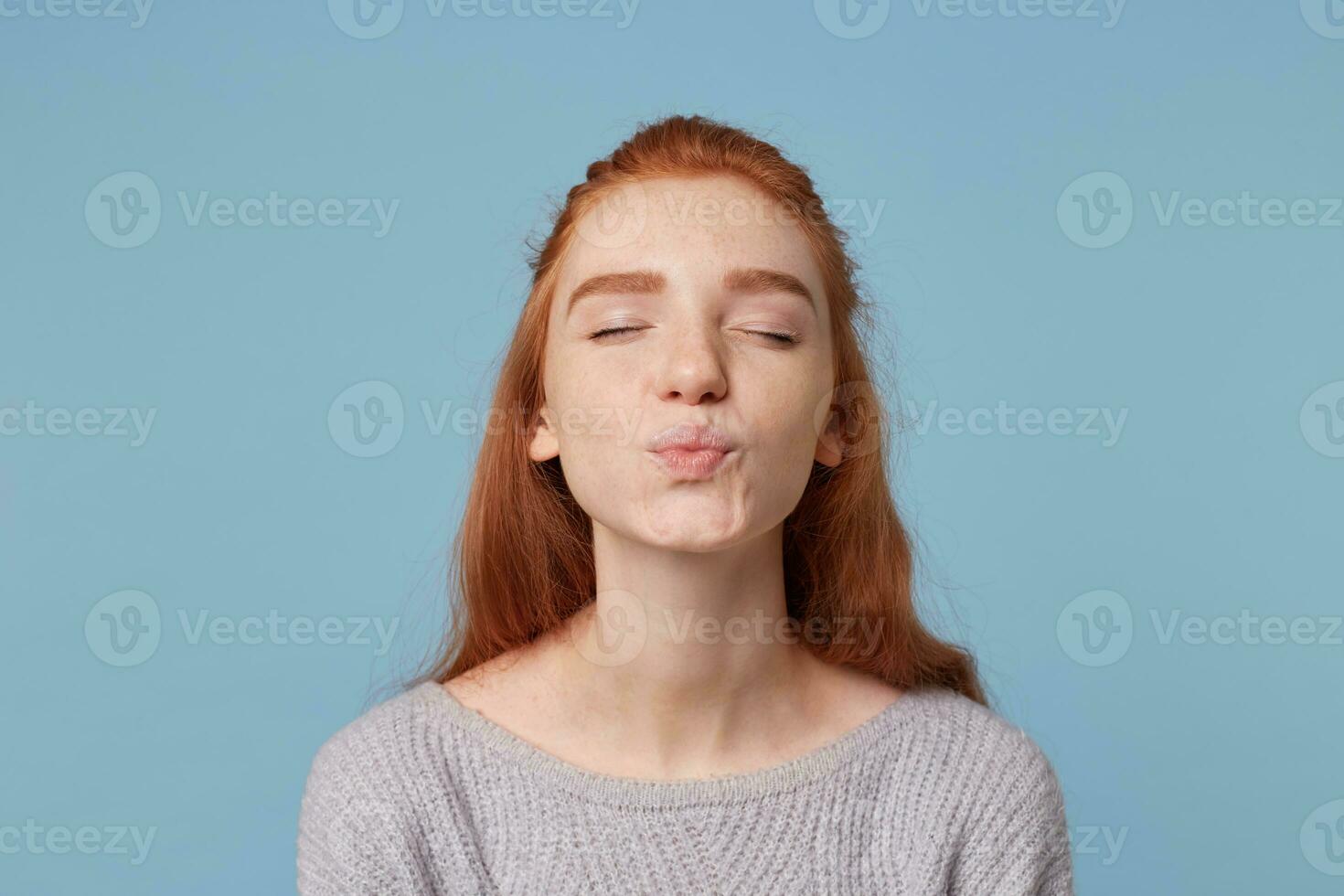ute nice young red-haired girl sends a kiss standing with eyes closed lips are folded for a kiss,over blue background. photo
