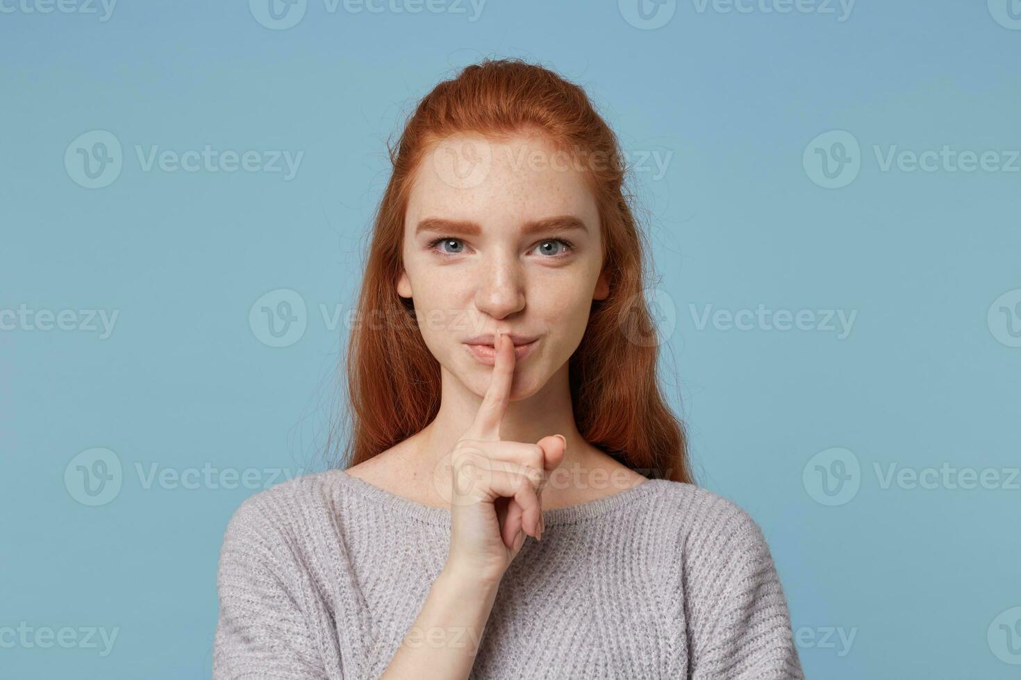 Portrait of nice cheerful red-haired young female depicts a gesture of silence,keeps finger at her mouth,jokingly looks at the camera, asks to keep a secret,mystery,over blue background. photo