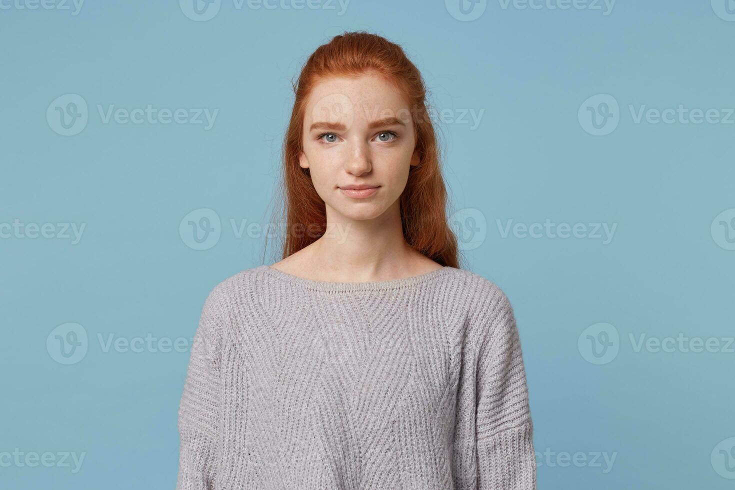 Young beautiful girl with long red hair and blue eyes with freckles looks straight into the camera feels calm and confidently isolated on a blue background photo