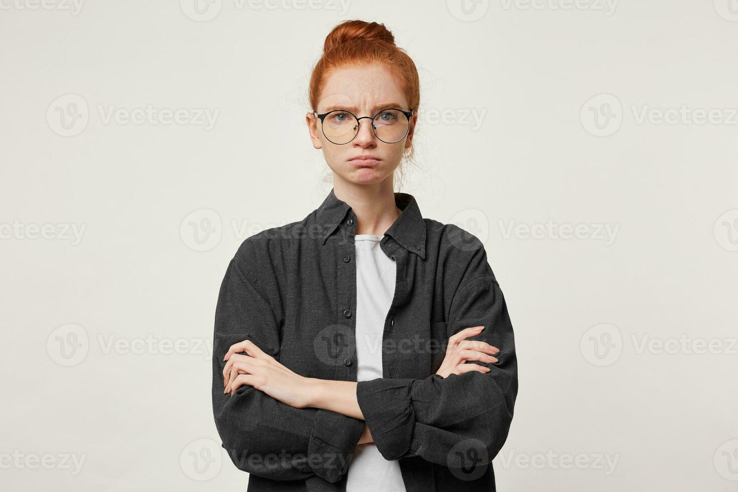 A young red-haired girl with bunched-up hair in glasses stands with her arms folded, dressed in a black men's shirt, looks strictly serious with frowned eyebrows, unfriendly facial expression. photo