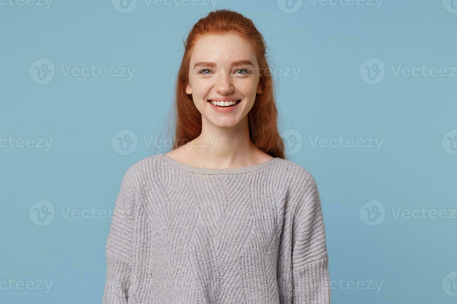 Portrait of a tender beautiful attractive young red-haired girl cute sincerely cheerfully affably smiling, showing her teeth, wearing an oversized warm sweater, isolated on a blue background photo