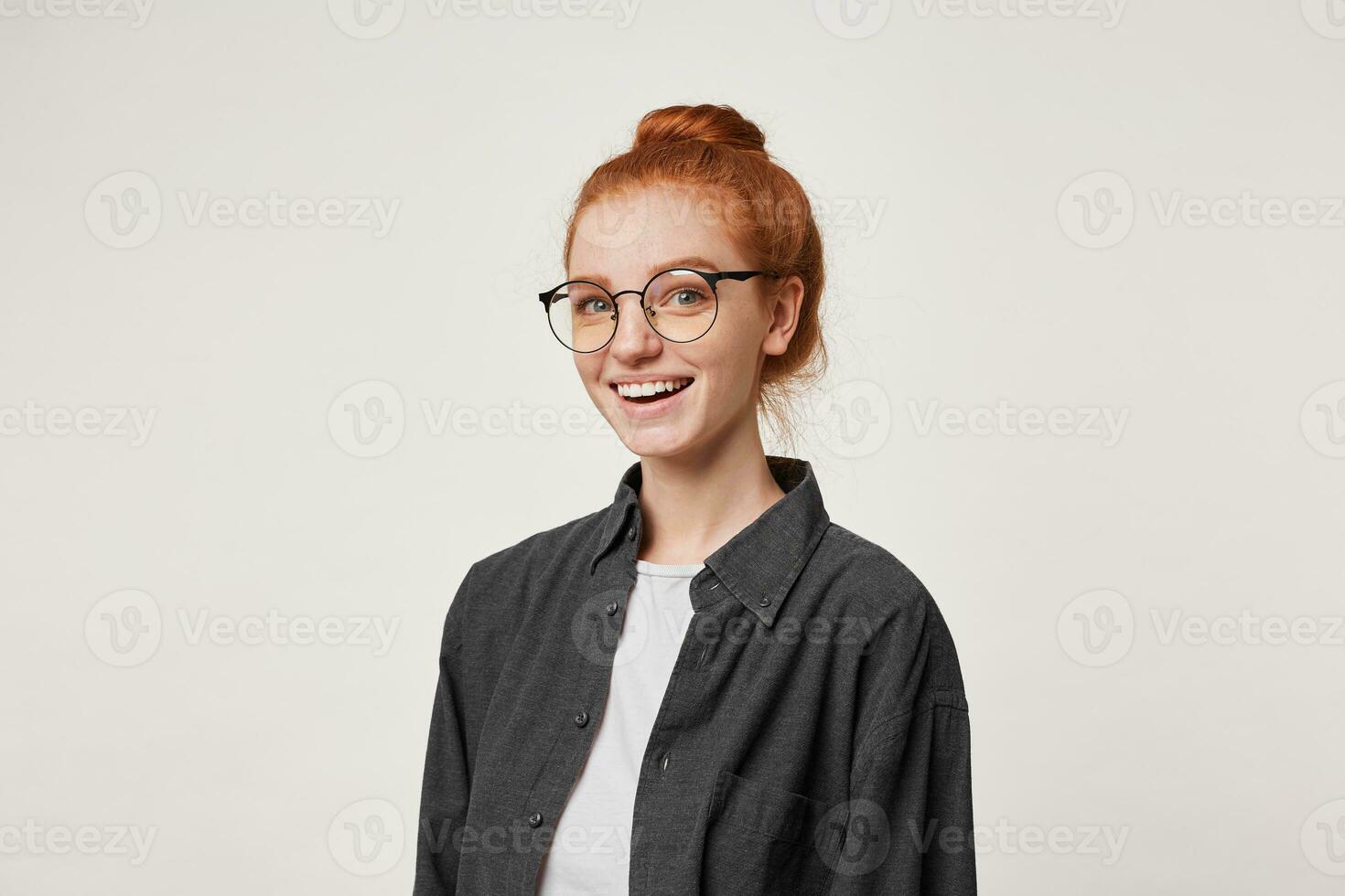 Cute red-haired girl looks into the camera through glasses, gently sincerely smiling, looks happy and satisfied, is half-turned, dressed in a black men's shirt, isolated on white background photo