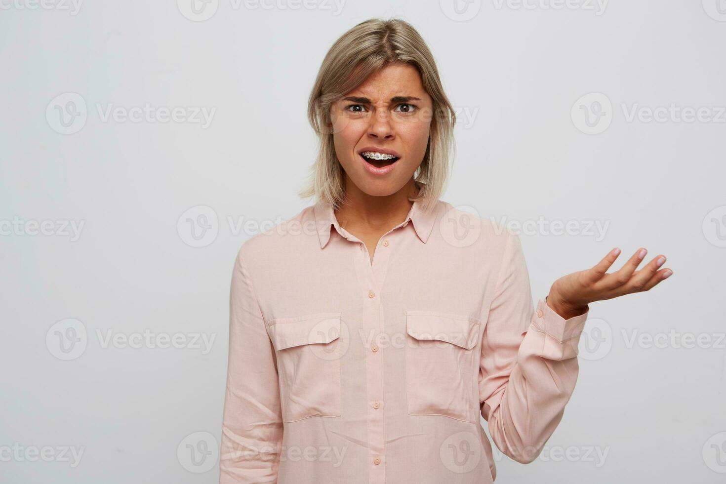 Closeup of displeased embarrassed blonde young woman with braces on teeth wears pink shirt looks confused and holding copy space on palm isolated over white background Feels unhappy photo