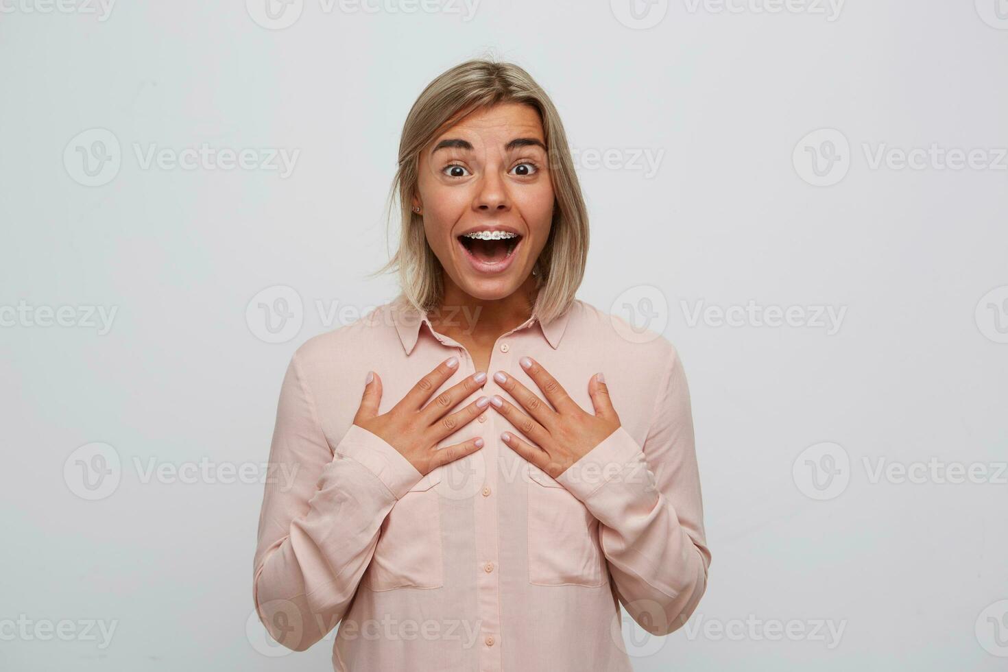 Portrait of surprised excited blonde young woman with opened mouth and braces on teeth wears pink shirt looks amazed and feels happy isolated over white background photo