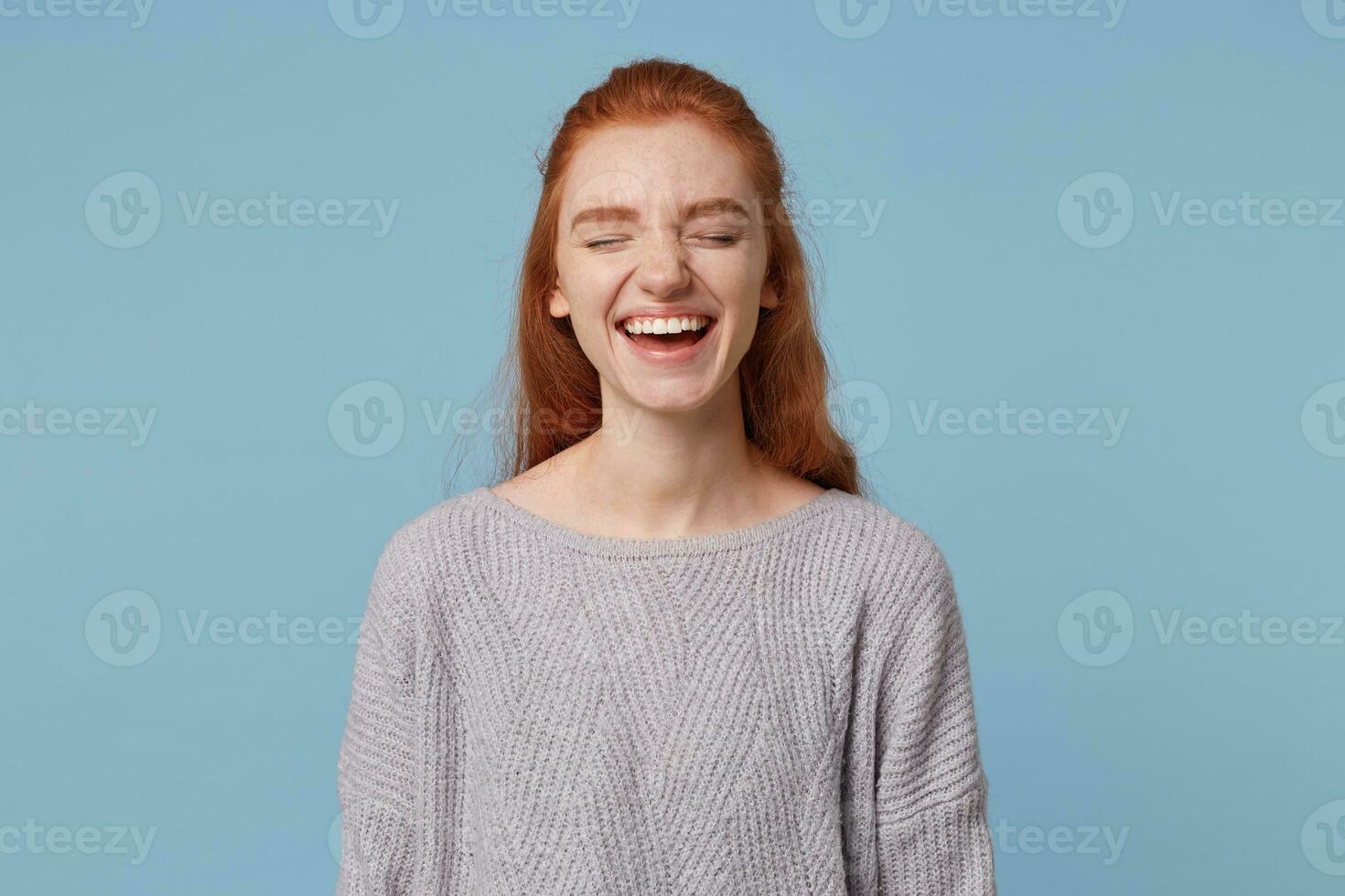 Portrait of a cheerful beautiful attractive young red-haired girl sincerely cheerfully laughing closing her eyes, wearing an oversized warm sweater, isolated on a blue background photo