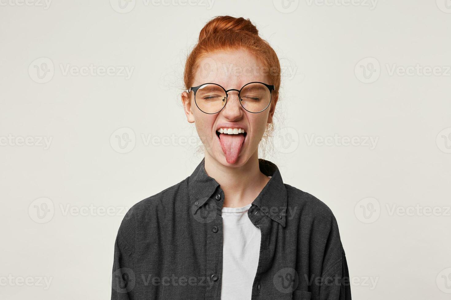 A young red-haired girl makes faces, sticks out shows her tongue, closes her eyes, wears a black man's shirt with glasses, has fun, plays photo