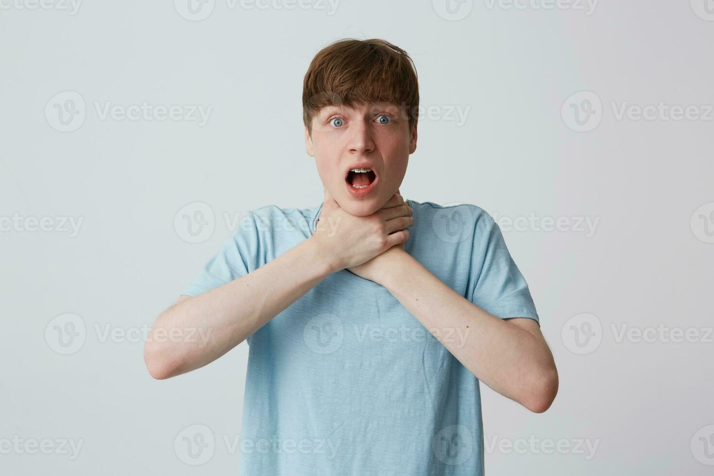 Closeup of despairing young man with braces on teeth wears blue t shirt have problems and strangles his neck with both hands isolated over white background photo