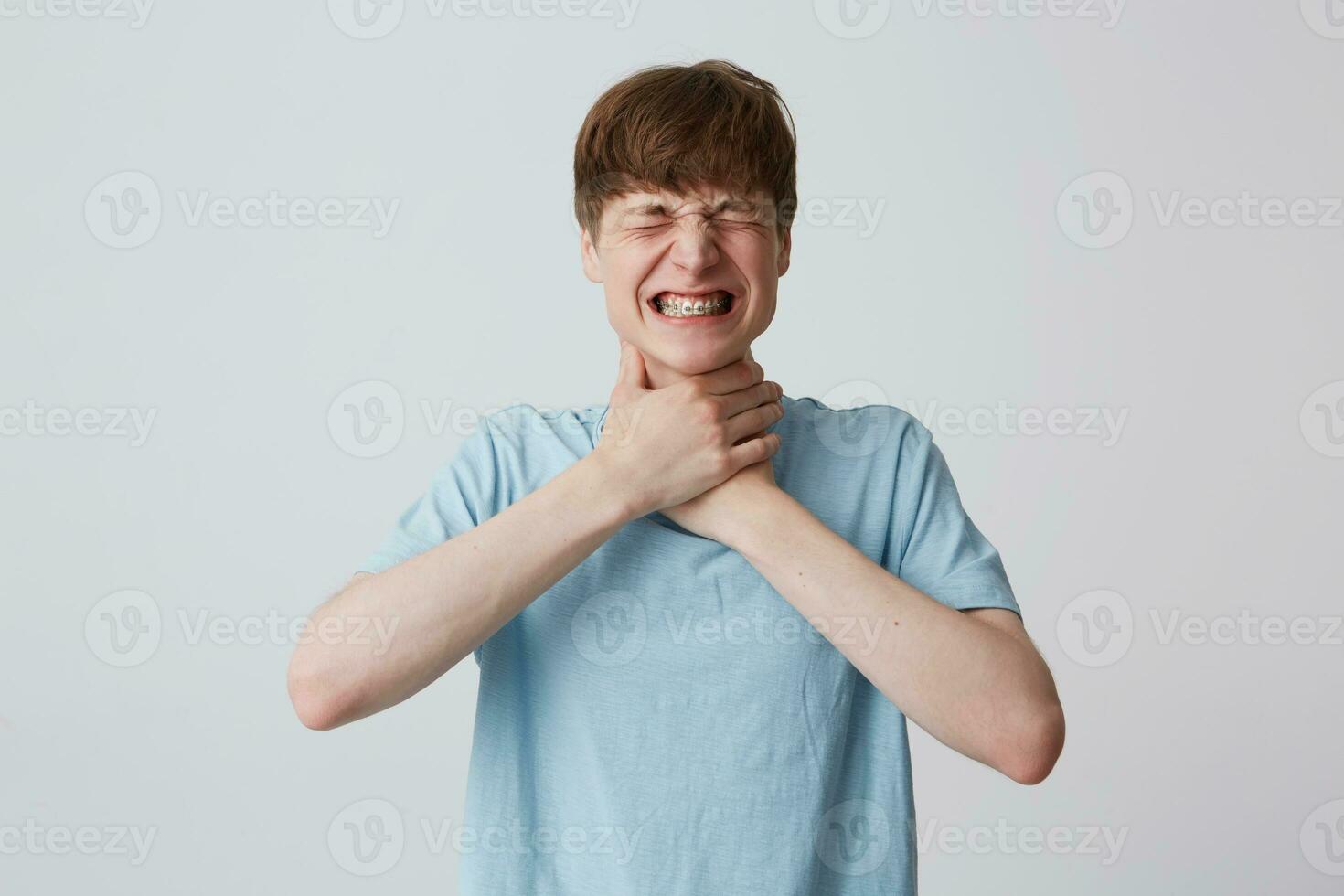 Portrait of hopeless stressed young man with closed eyes and braces on teeth wears blue t shirt strangles himself with both hands isolated over white background photo