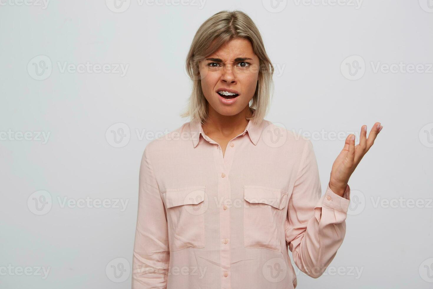 Portrait of unhappy confused young woman with blond hair and braces on teeth wears pink shirt feels embarrassed and holding copy space on palm isolated over white background photo