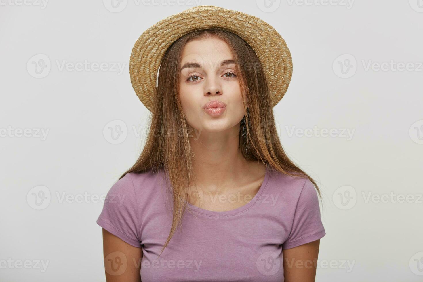 Close up of cute lovely girl in casual outfit in a straw hat with a pink ribbon sending blowing kiss with pout lips looking at camera isolated on white background. Affection feelings concept photo