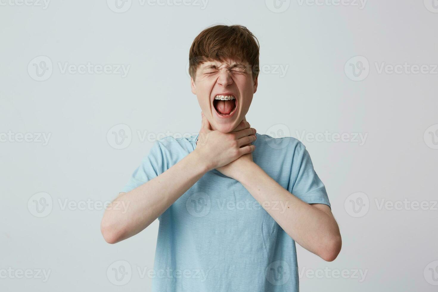 Portrait of crazy desperate young man student with closed eyes and braces on teeth wears blue t shirt strangles himself with both hands and screaming isolated over white background photo