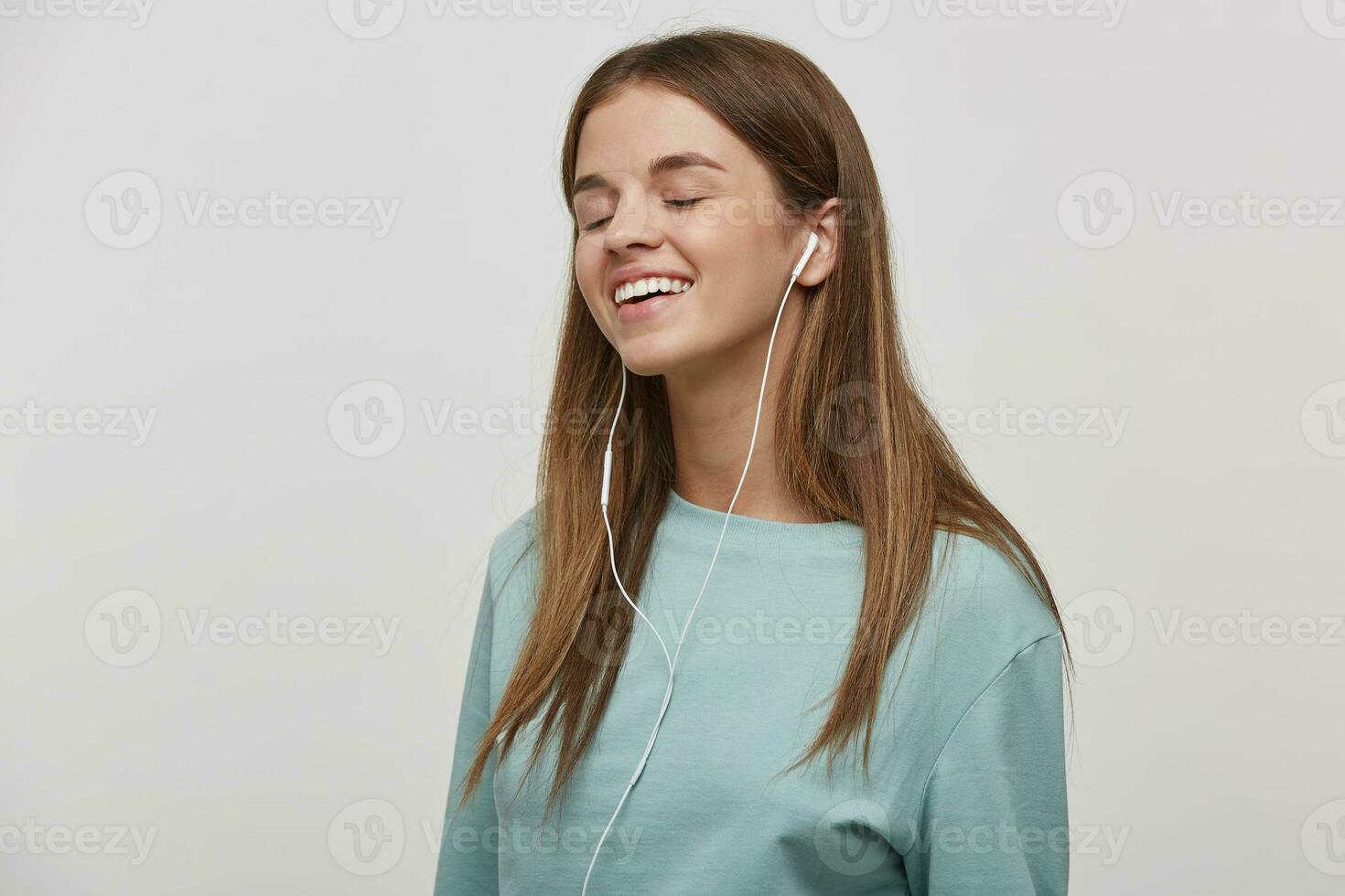 afilado en música joven chica, sonrisas, escucha favorito música con auriculares, soportes oblicuo, con cerrado ojos, vestido casual, en un blanco antecedentes en el estudio. foto