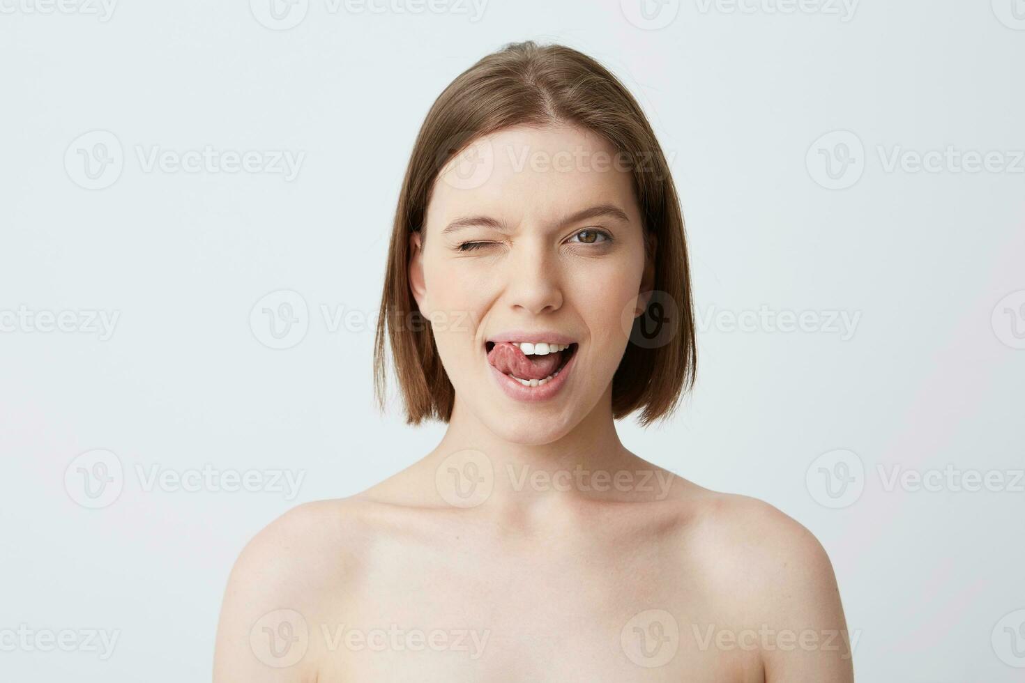 Close up shot of cute playful young woman with dark hair and perfect skin shows tongue and winks isolated over white background Standing half naked after shower photo