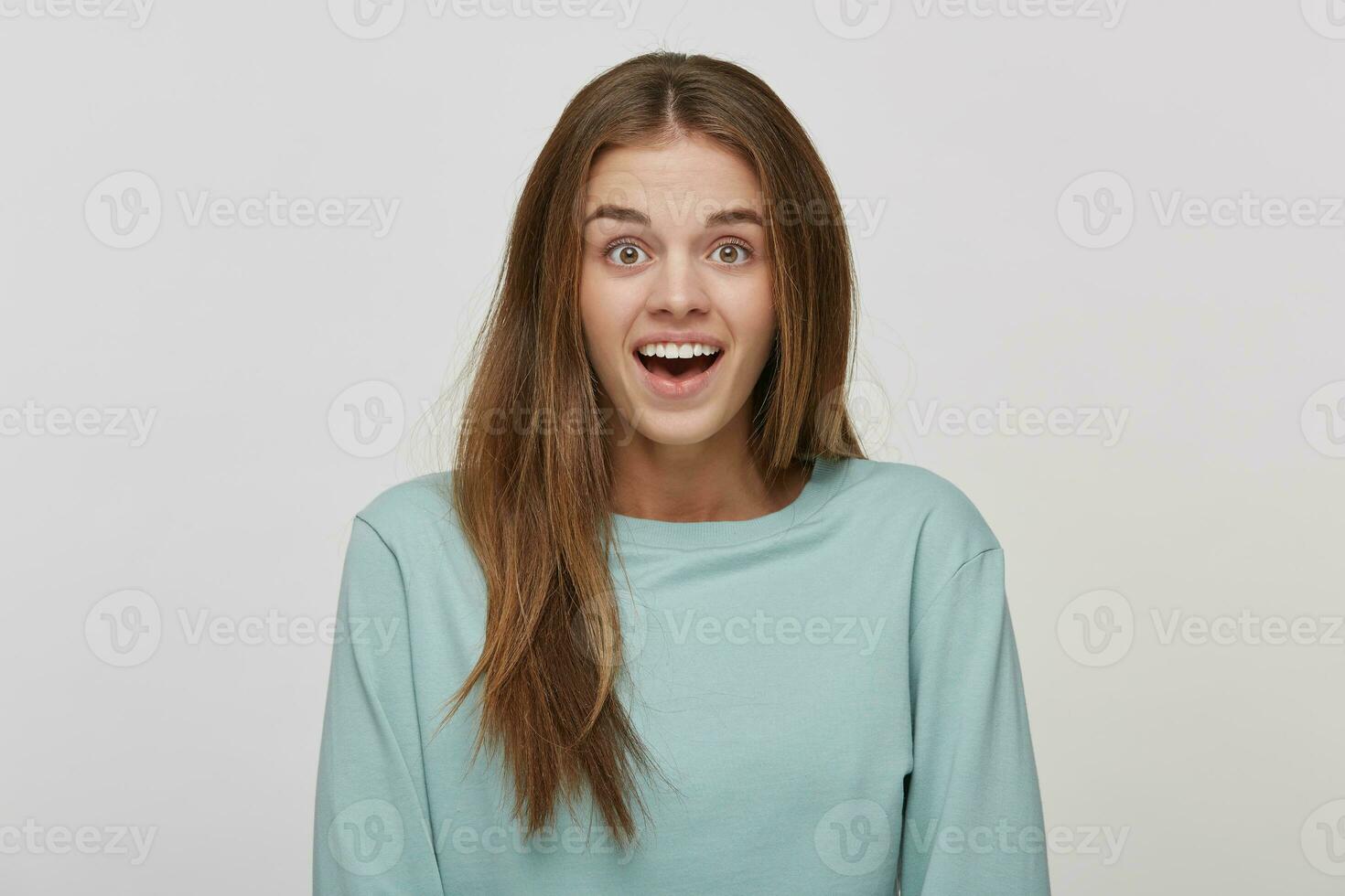 sorprendido alegre hermosa hembra con asombrado expresión, mira con molestado ojos y mantiene boca abierto, vestido casual azul largo manga camiseta, terminado blanco antecedentes. humano reacción y emociones foto