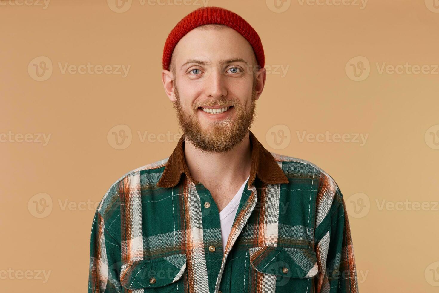 Happy cheerful young man wearing red hat and shirt rejoicing at positive news, looking at camera with joyful and charming smile. Student guy relaxing indoors after college photo