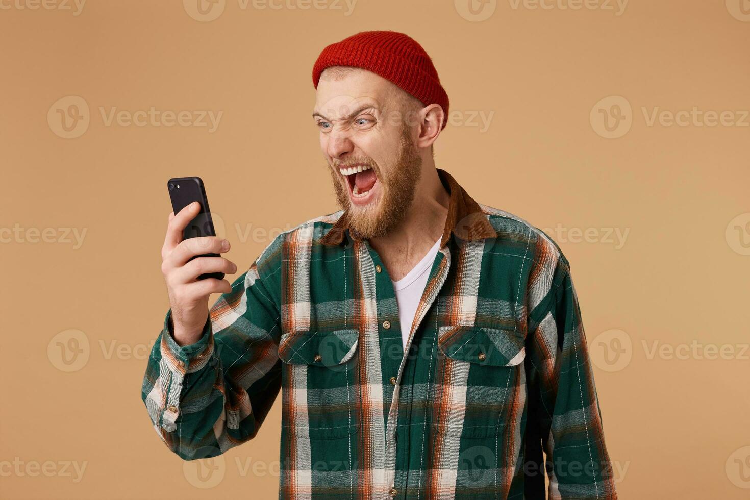 Portrait of angry young man screaming on his mobile phone over beige background. Mad irritated bearded young man in plaid shirt holding and shouting on cell phone photo