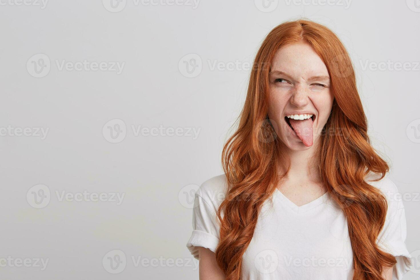 Portrait of cheerful playful young woman with long wavy red hair and freckles wears t shirt winks and shows tongue isolated over white background photo