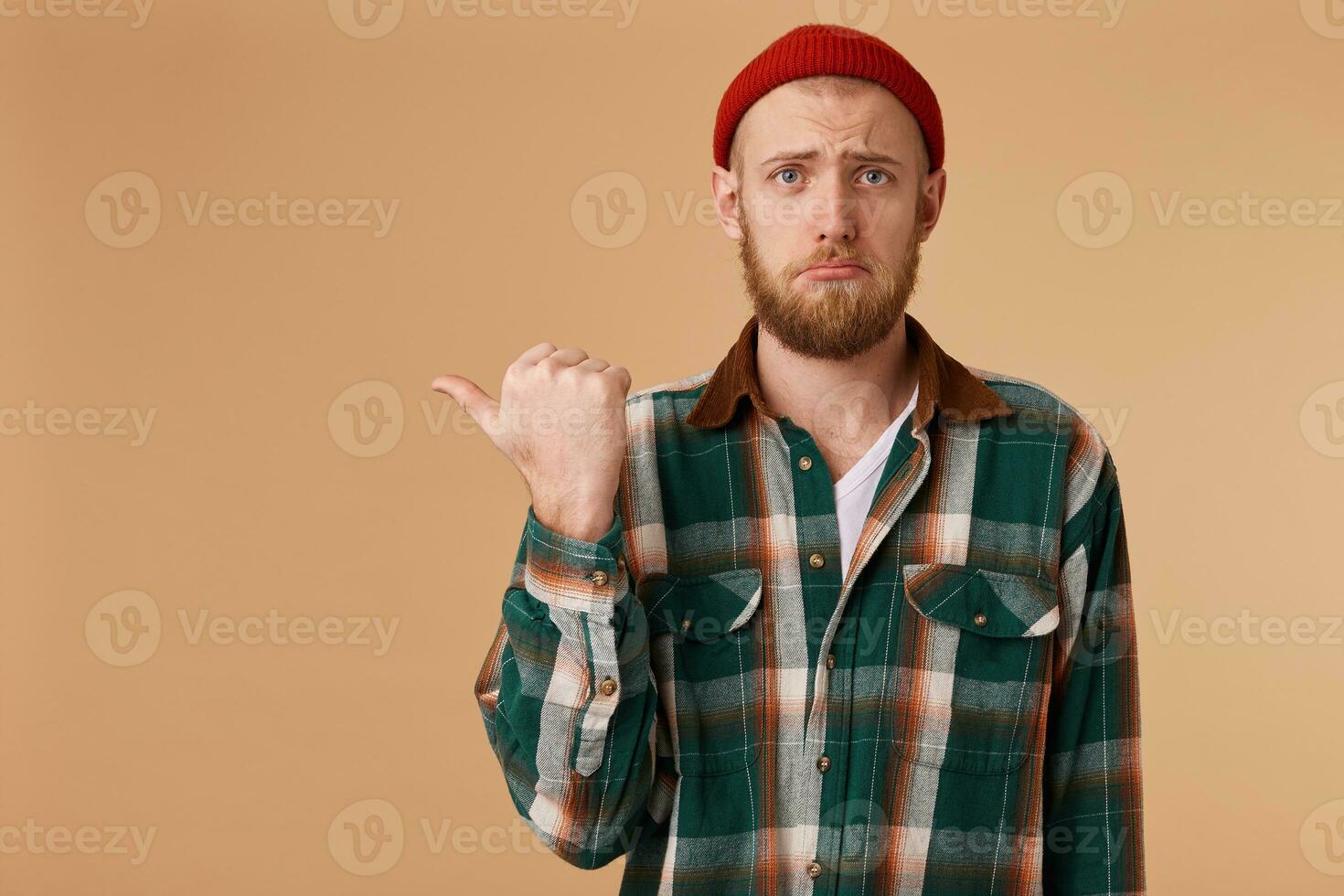 An upset guy with a sad look in his eyes with a displeased facial expression shows his thumb on his left hand on the empty space for promotion. photo