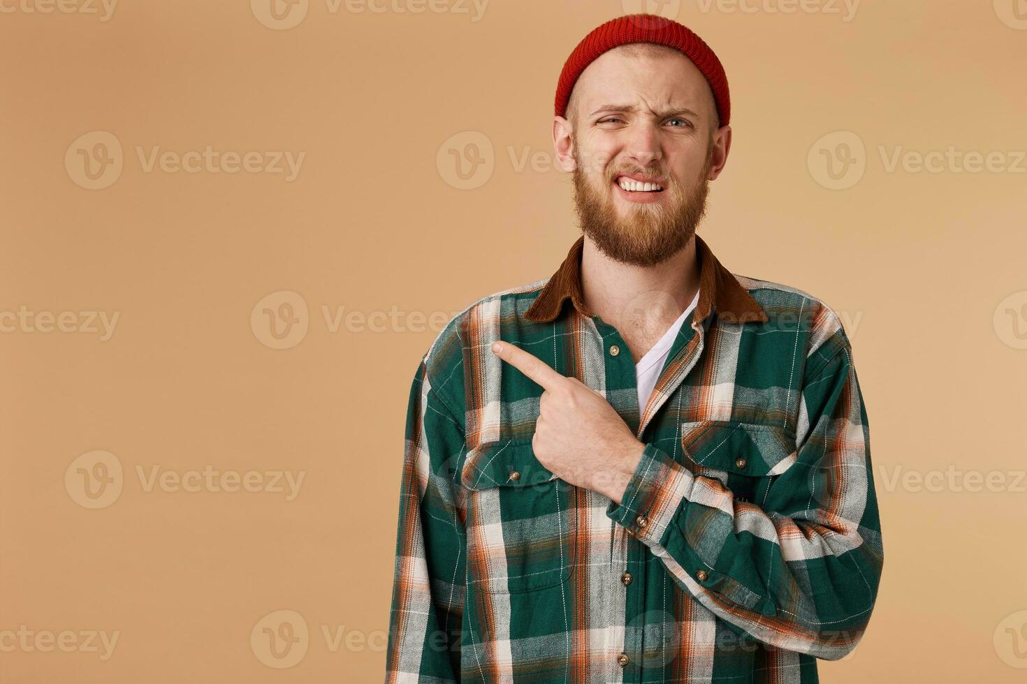 Unhappy bearded male in red hat without pleasure points with index finger at left corner, shows copy space for advertisement or promotional text isolated over beige background photo